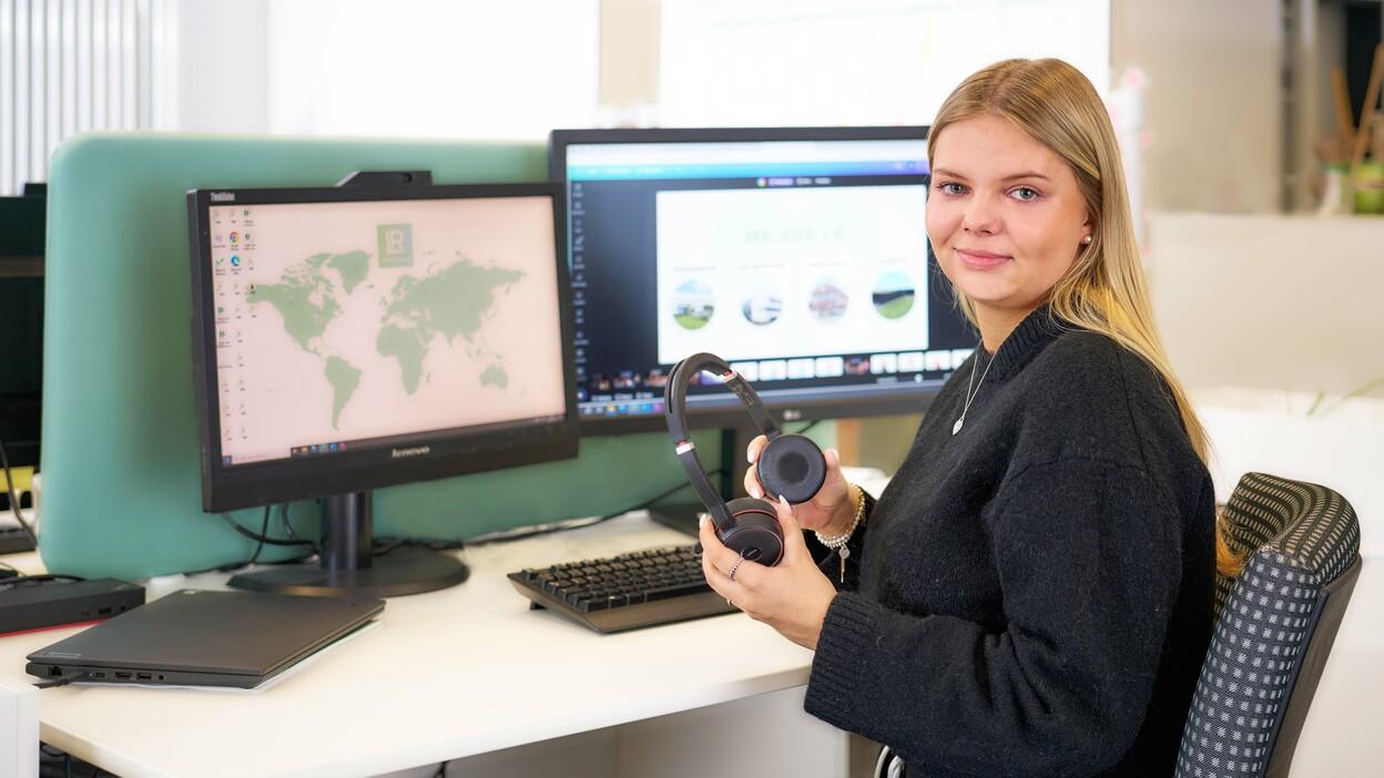 Auszubildende an einem Arbeitsplatz mit zwei Monitoren und Headset in der Hand.